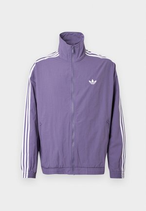 Fioletowa kurtka na zamek z wysokim kołnierzem, białe trzy paski na rękawach i małe białe logo na lewej piersi. Materiał: poliester.