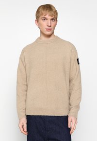 Maglione beige lavorato a maglia con scollo rotondo, orlo a coste e un tessuto testurizzato. Presenta una patch con logo nera sulla manica sinistra.
