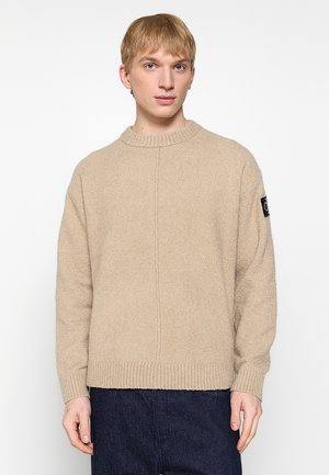 Beige knitted sweater with a round neckline, ribbed hem, and a textured fabric. Features a black logo patch on the left sleeve.