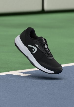 Black athletic shoe with white sole and white logo hovering above blue and green tennis court surface.