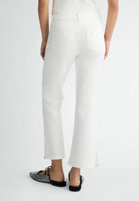 Weiße, ausgestellte Jeans mit Taschen auf der Rückseite und seitlichen Schlitzen am Saum. Kombiniert mit schwarzen, verzierten Mules mit spitzen Zehen. Glatte Textur.