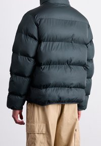 Ciemnozielona kurtka puchowa o pudełkowym kroju, z pikowanymi sekcjami i wysokim kołnierzem. Wykonana z gładkiego, błyszczącego materiału.