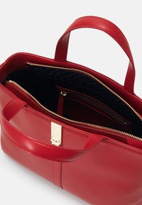 Sac à main en cuir rouge avec deux poignées, un fermoir doré et une fermeture éclair supérieure partiellement ouverte montrant un intérieur noir avec une poche intérieure zippée.