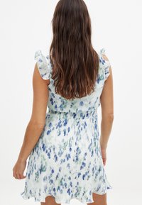 Robe florale avec des épaules à volants, fabriquée à partir d'un tissu léger. Présente des motifs de fleurs bleues et vertes sur fond blanc. Courte et plissée.