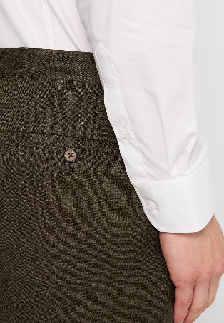 Witte lange mouwen shirt met knopenmanchetten, gedragen met donkerbruine textuurbroek met een achterzak en knoopdetail.