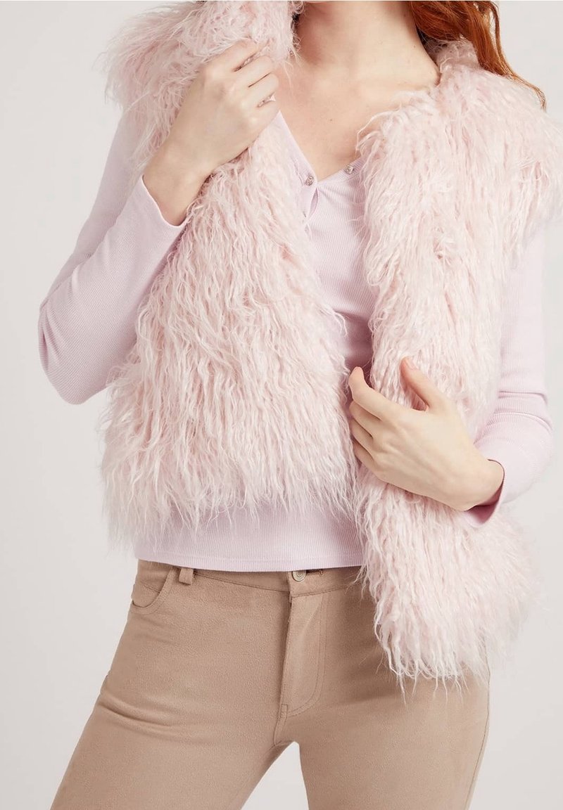 Femme portant un haut à manches longues rose clair et un pantalon beige, tenant une veste en fourrure rose pâle et duveteuse autour de ses épaules.
