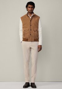 Brauner Wildlederwesten mit zwei Vordertaschen, Knopfverschlüssen und einem Kragen, über einem weißen Langarmshirt und beigen Hosen getragen. Schwarze Stiefel.