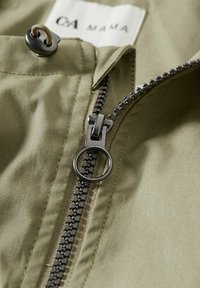 Gros plan d’une veste en tissu kaki partiellement dézippée avec une fermeture éclair métallique et un tirage rond près d’un bouton et d’une étiquette en tissu.