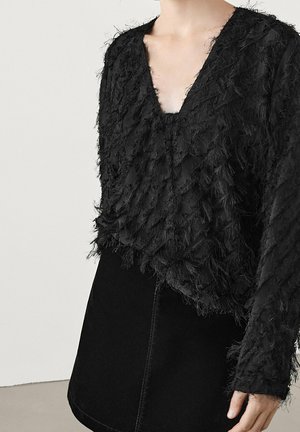 Czarny teksturowany top z długim rękawem i dekoltem w serek, zestawiony z gładką czarną spódnicą, widoczny od połowy tułowia do górnej części uda.