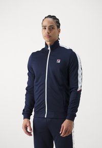 Marineblauwe zip-up trainingsjack met witte zijstrepen, hoge kraag en Fila-logo op de borst. Zacht, rekbaar materiaal, aansluitend ontwerp.