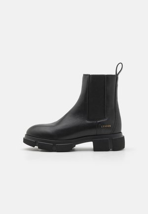 Černá kožená kotníková bota s elastickými bočními panely, robustní gumovou podrážkou a kontrastním logo detailem na patě. Hladká textura, zaoblená špička.