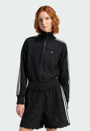 Donna dai capelli rossi che indossa una giacca nera con zip, decorata da motivi, e pantaloncini abbinati con strisce bianche sulle maniche e sui lati.