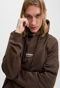 Νεαρό άτομο με μακριά ξανθά μαλλιά που φοράει ένα καφέ φούτερ JJ Studios, ακουμπώντας το χέρι του με το τατουάζ "SPACE IDOL" κοντά στο πρόσωπο.