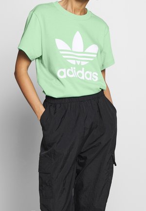 Person iført lysegrøn Adidas t-shirt med hvidt logo og sorte løse cargobukser, hænderne i lommerne, mod ensfarvet grå baggrund.
