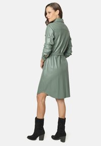 Vestido verde de cuero sintético con cuello de camisa, mangas largas, cintura ceñida con cinturón y dobladillo curvado. Combinado con botas negras hasta el tobillo.