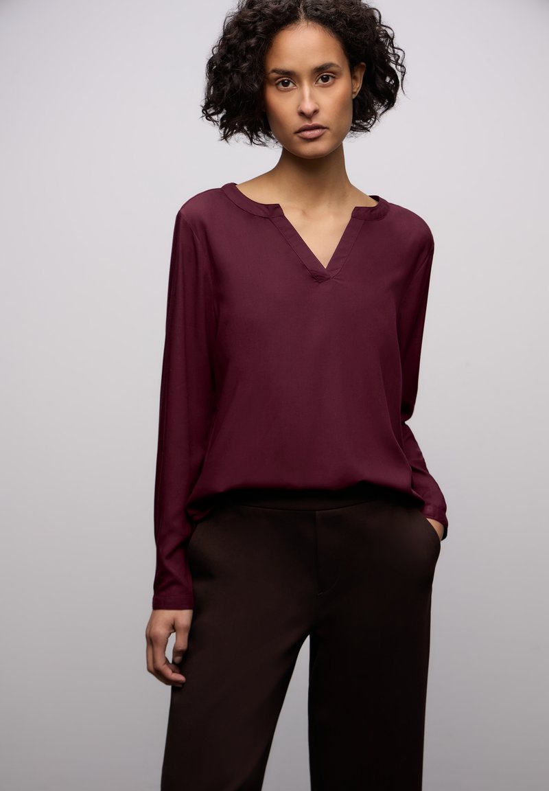 Bordeaux langærmet bluse med V-udskæring, lavet af glat stof, parret med slim-fit brune bukser, der har sidelommer.