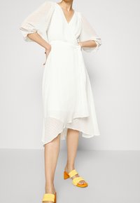 Vit omslagsklänning med prickig textur, korta genomskinliga puffärmar och asymmetrisk nederkant. I kombination med gula blockklack-sandaletter.