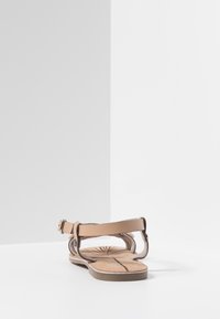 Sandal plano beige con tiras en el tobillo, cierre con hebilla y diseño de hojas en la plantilla, fotografiado contra un fondo blanco en la esquina.