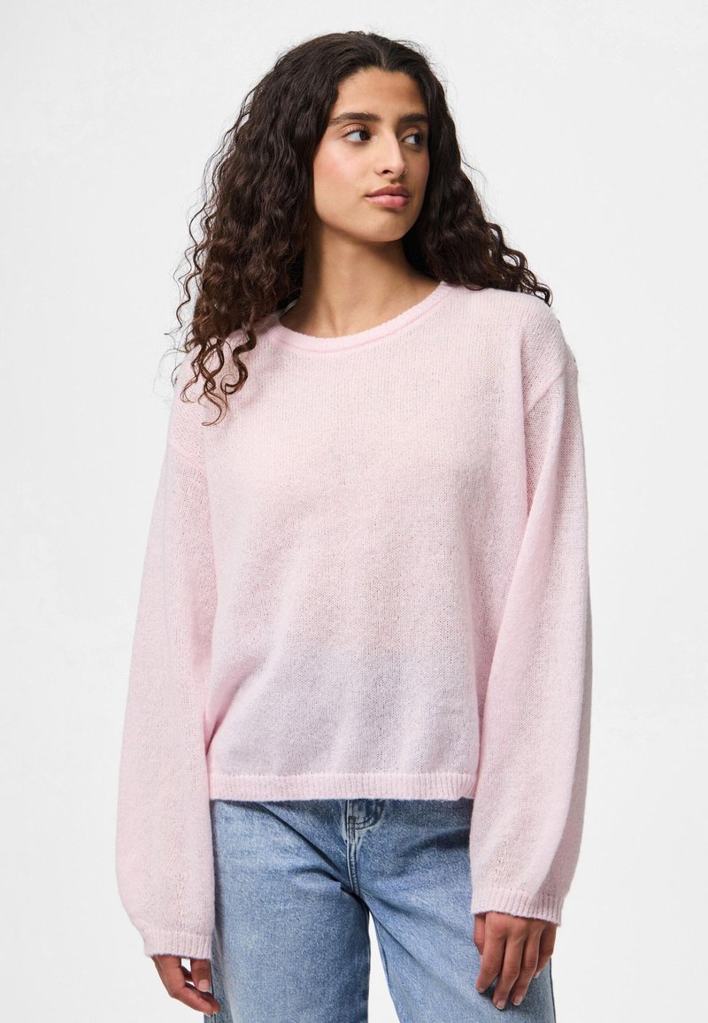Woman with long curly hair wearing a light pink sweater and blue jeans, looking slightly to her left against a plain white background.
