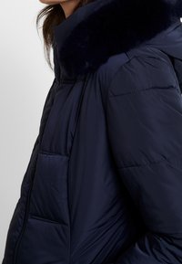 Navy puffer jacket featuring a fur-lined hood, quilted texture, side pockets, and a zip closure. Smooth material with a sleek design.