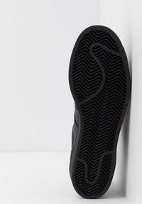 Svart sneaker-sula med strukturerat zigzag- och böljemönster, placerad vertikalt mot en vit hörnbakgrund.