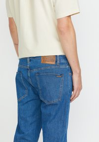 Jeans in denim blu con gamba dritta, dotati di una toppa in pelle marrone con logo, due tasche posteriori e cuciture in evidenza.