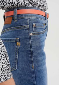 Modré džíny z denimu s vysokým pasem, vyztuženým šitím, zlatými akcenty a červeným koženým páskem, se zaobleným logem na zadní straně.