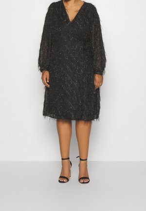 Kvinde iført knælængde sort slå-om-kjole i gennemsigtigt, tekstureret stof med lange ærmer, kombineret med sorte åbne ankelstrop-sandaler.