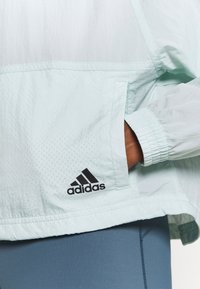 Handen i fickan på en ljus mintgrön Adidas-jacka som bärs över blå leggings, med Adidas-logotypen synlig på jackans framsida.