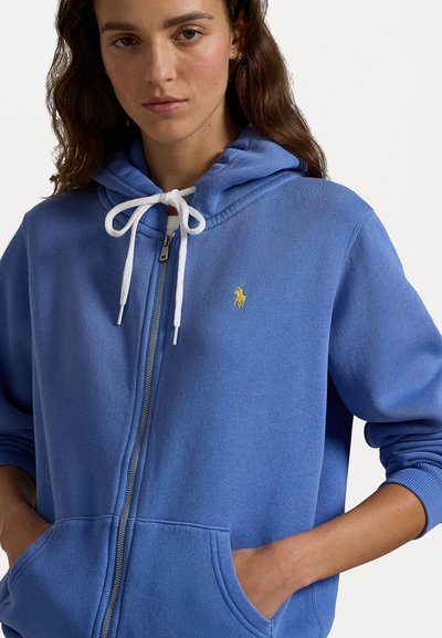 Blaues Zip-Hoodie mit weißen Kordeln, großen Vorderaschen und einem kleinen gelben Logo auf der Brust. Weicher Stoff und lockere Passform.