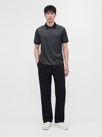 Homme debout les mains dans les poches, portant un polo noir à manches courtes à motifs, un pantalon noir et des baskets blanches sur un fond uni.