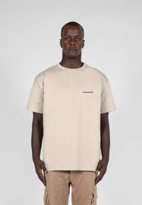 Tricou din bumbac bej, cu guler rotund și mâneci scurte; are un logo negru pe pieptul stâng. Purtați cu pantaloni cargo bej.