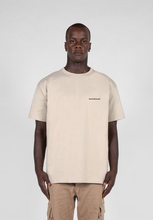 Beige cotton t-shirt with crew neck and short sleeves; features a black logo on the left chest. Worn with tan cargo pants.