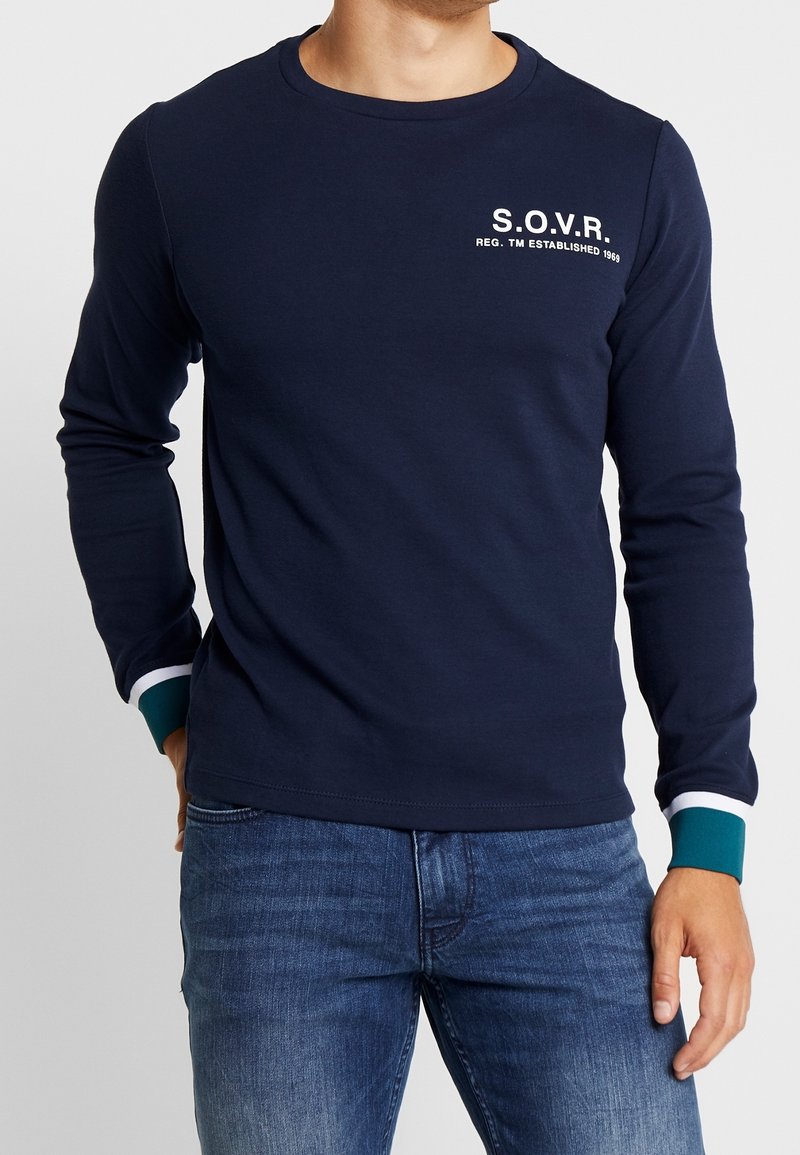 Mężczyzna w granatowej koszuli z długim rękawem z napisem "S.O.V.R." na klatce piersiowej, z kontrastującymi mankietami w kolorze turkusowym i białym, w połączeniu z niebieskimi dżinsami.