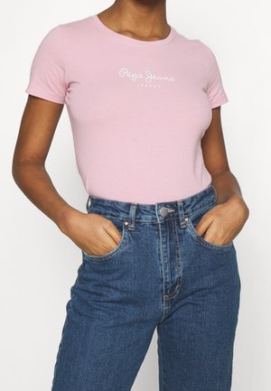 Kobieta ubrana w jasnoróżowy t-shirt Pepe Jeans London włożony do wysokich niebieskich dżinsów, ręce w kieszeniach.