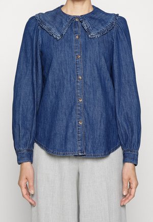 Vrouw draagt een blauw denimblouse met lange mouwen, gerimpelde Peter Pan-kraag en knoopsluiting aan de voorkant, gecombineerd met lichtgrijze broek.