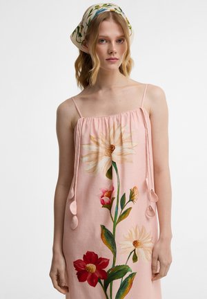 Fiatal nő rózsaszín virágos ruhát visel vékony pántokkal és virágos fejkendővel, egyszerű fehér háttér előtt áll.