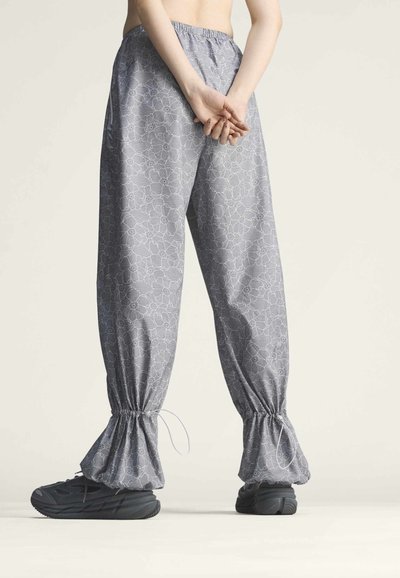 Personne portant un pantalon ample gris à motif floral avec poignets élastiques et cordon, debout, les mains jointes derrière le dos et chaussures noires.