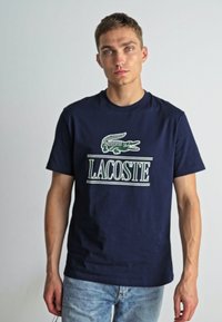 Camiseta de algodón en color azul marino con un logo de cocodrilo verde y el texto "LACOSTE". Mangas cortas, cuello redondo y un corte relajado.