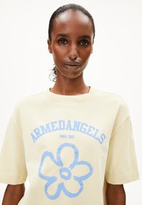 Helles gelbes T-Shirt mit blauem Blumenmotiv und dem Text "ARMEDANGELS". Kurze Ärmel und Rundhalsausschnitt. Weiches Baumwollmaterial.