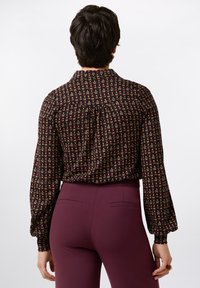 Schwarze Bluse mit buntem Blumenmuster, langen Ärmeln und lockerer Passform, kombiniert mit bordeauxroten, hochgeschnittenen Hosen mit Gesäßtaschen.