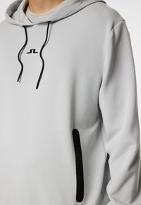 Lys grå hoodie med et sort logo og snore. Har en side-lomme med lynlås i sort, lavet af glat, blødt materiale.