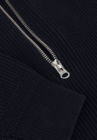 Nahaufnahme eines teilweise geöffneten silbernen Reißverschlusses auf schwarzem geripptem Strickstoff mit einem gefalteten Ärmel.