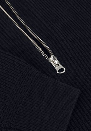 Nahaufnahme eines teilweise geöffneten silbernen Reißverschlusses auf schwarzem geripptem Strickstoff mit einem gefalteten Ärmel.