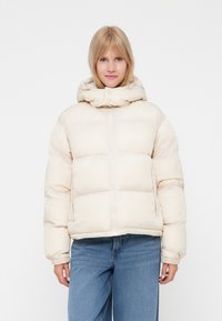 Puffjacka i krämfärg, med hög krage, dragkedja fram och elastiska ärmslut. Designen inkluderar sömnadsdetaljer för isolering.
