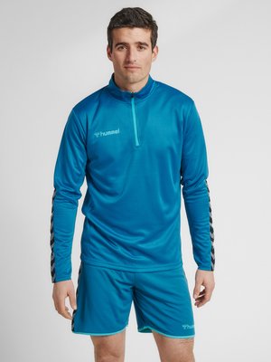 Uomo con top sportivo blu abbinato con zip a quarto e pantaloncini, in piedi davanti a uno sfondo chiaro e semplice.