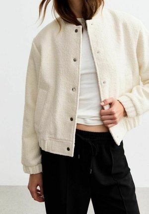 Kobieta ubrana w kremową kurtkę polarową na guziki, na białym prążkowanym topie i czarnych spodniach z troczkiem, z jedną ręką regulującą kurtkę.