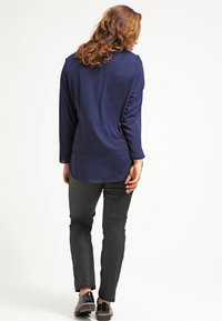 Blusa a maniche lunghe blu navy con vestibilità ampia, abbinata a pantaloni neri slim-fit e scarpe nere. Il tessuto appare liscio e leggero.