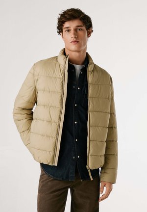 Beige gewatteerde jas met een hoge kraag, voorzien van gestikte panelen en een rits aan de voorkant. Draag deze over een donkerdenim shirt en bruine broek.