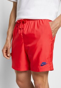 Muž v jasně červených šortkách Nike s elastickým pasem a šňůrkou, ruka v kapse, viditelný lem bílého trička.